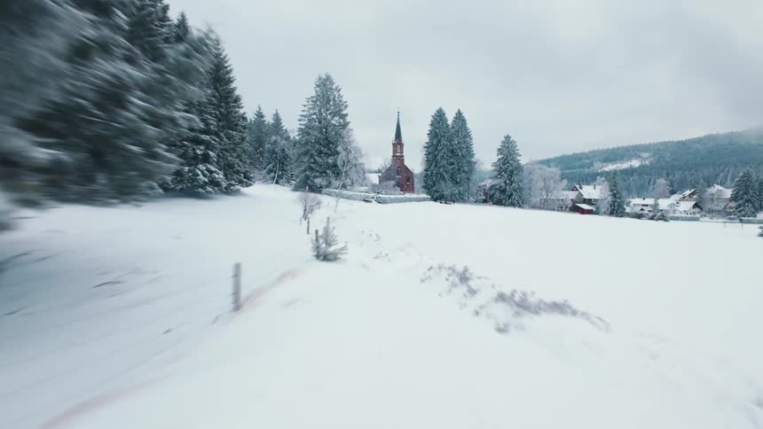 A camera moves quickly over a thick layer of white snow toward a brick church with a tall steeple surrounded by frost covered evergreen trees and small houses under a cloudy winter sky