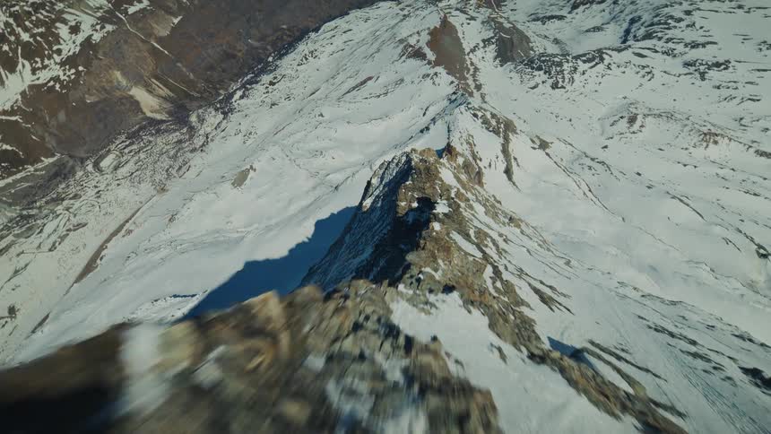 A fast aerial view descends over a jagged rocky mountain peak and its sharp ridge covered in white snow under a bright clear sky in a winter landscape