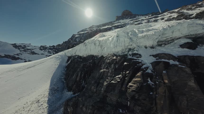 A high angle drone camera pans upward along a jagged rocky cliffside covered in thick glacial ice and white snow toward a sharp mountain peak under a bright sun and clear blue sky