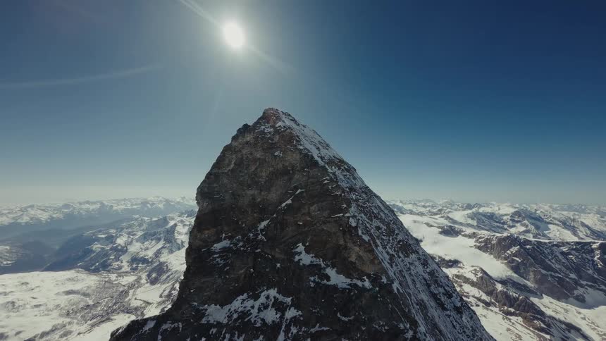 Snow on the rough terrain of a mountain peak with mountains in the distance as the sun shines brightly overhead