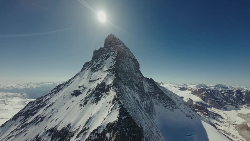 The Matterhorn stands under a bright sun and clear blue sky while rocky ridges and smaller frozen ranges stretch across the distant horizon