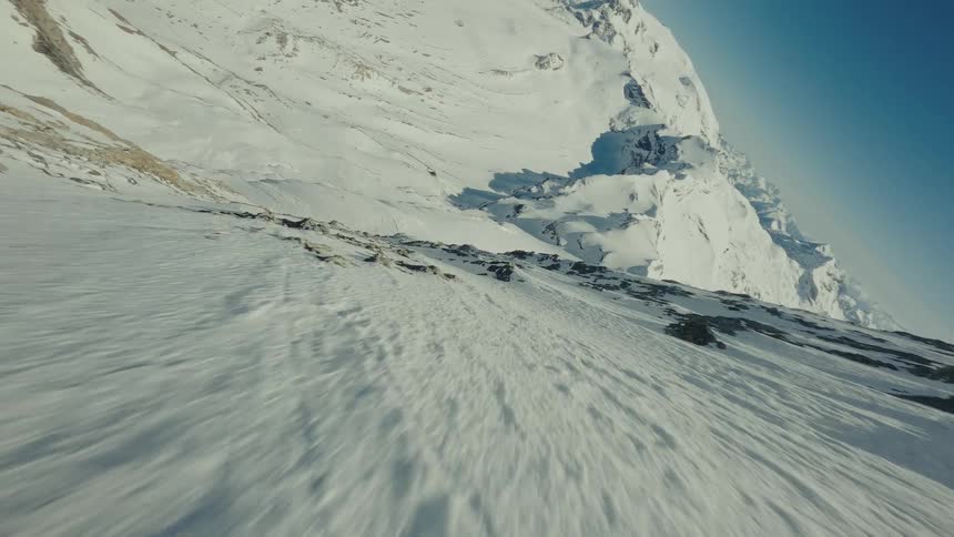 A fast moving camera flies alongside a steep mountain slope covered in white snow and dark rocks while a vast winter range of snowy peaks stretches into the distance under a clear blue sky