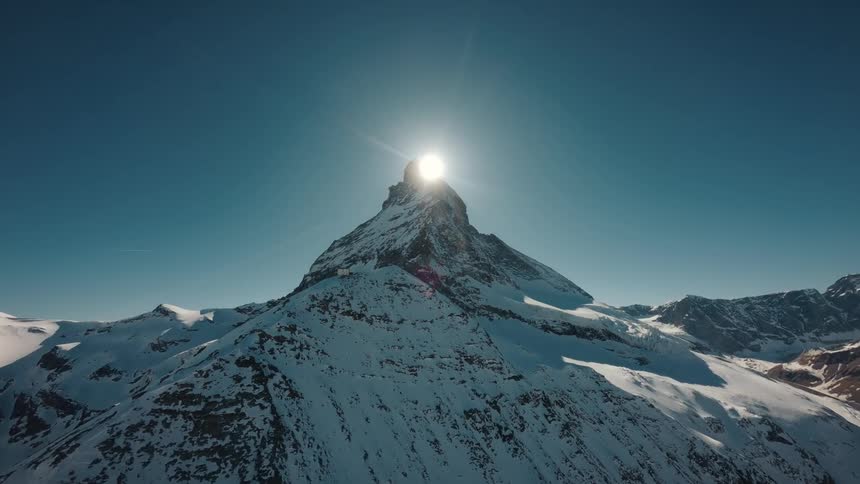 A wide view of a pointed mountain peak covered in snow as the bright sun rises directly behind the summit against a clear blue sky in a winter landscape