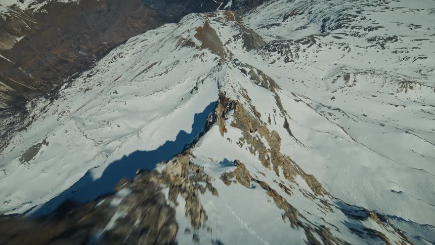 A fast aerial view glides over a sharp rocky mountain ridge covered in patches of white snow under a bright clear sky in a cold winter environment