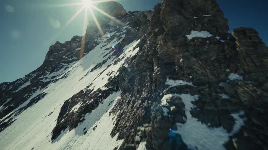 A fast moving camera travels up a steep rocky mountain face towards a sharp peak where the bright sun creates a brilliant flare against a clear blue sky