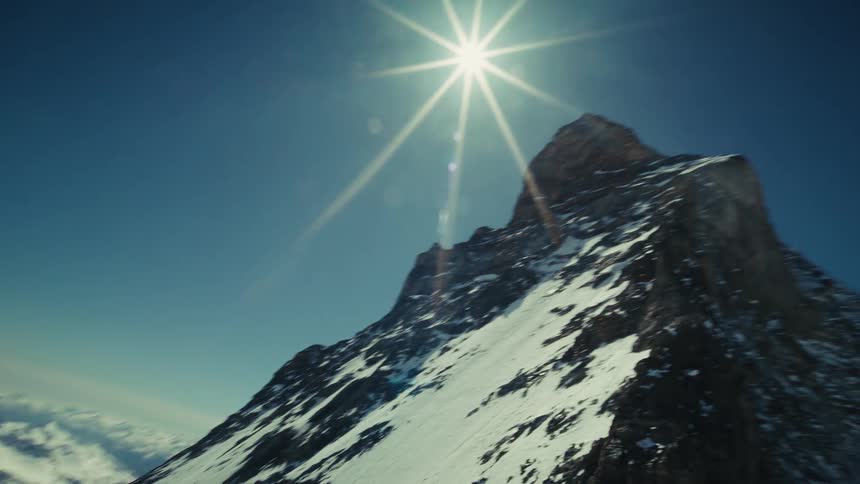 An aerial camera flies over a sharp snowy ridge before tilting up to reveal the sun shining directly above the jagged peak of a massive mountain