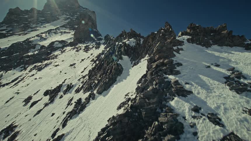 A fast moving camera ascends the steep rocky slopes of the matterhorn in the swiss alps where jagged dark stones and white snow patches meet under a bright sun and clear blue sky
