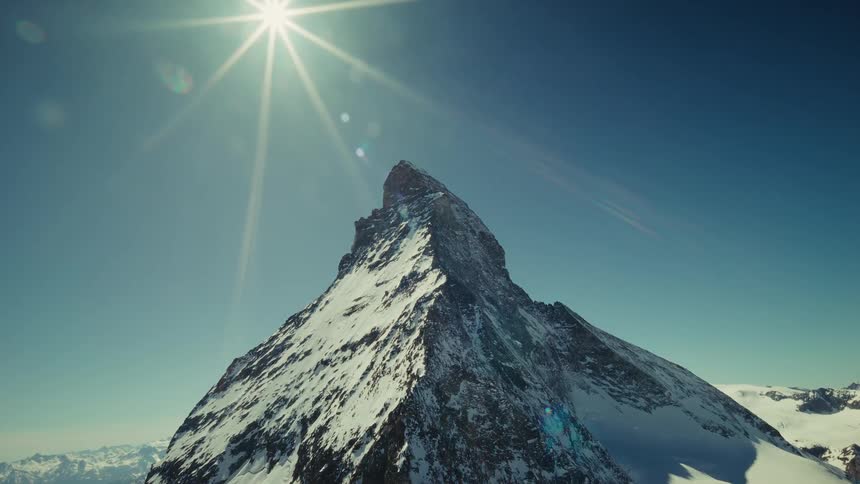 A high view of the matterhorn covered in snow and ice stands against a deep blue sky with the bright sun creating a lens flare in the upper left corner of the frame