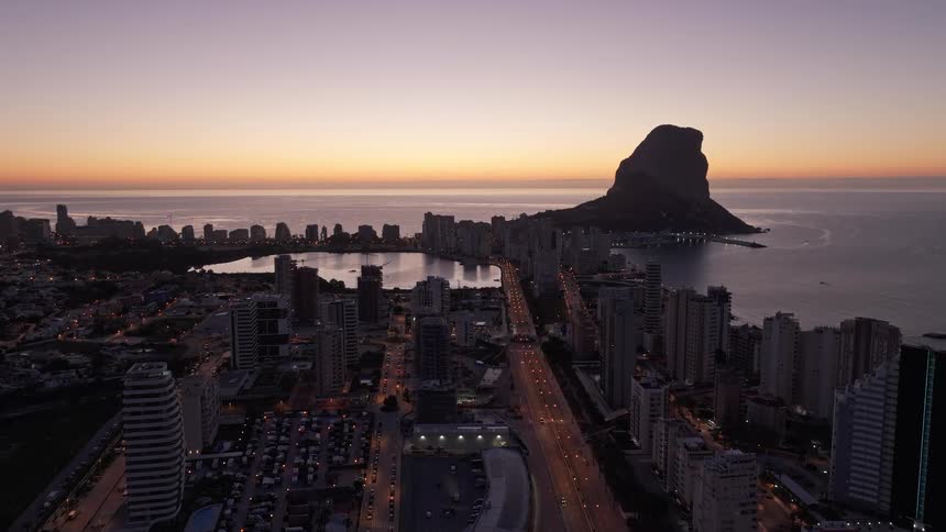 A wide aerial view shows the city of calpe at dawn with the peon de ifach landmark rising over the mediterranean sea as orange light glows on the horizon and streetlights illuminate the roads between tall buildings