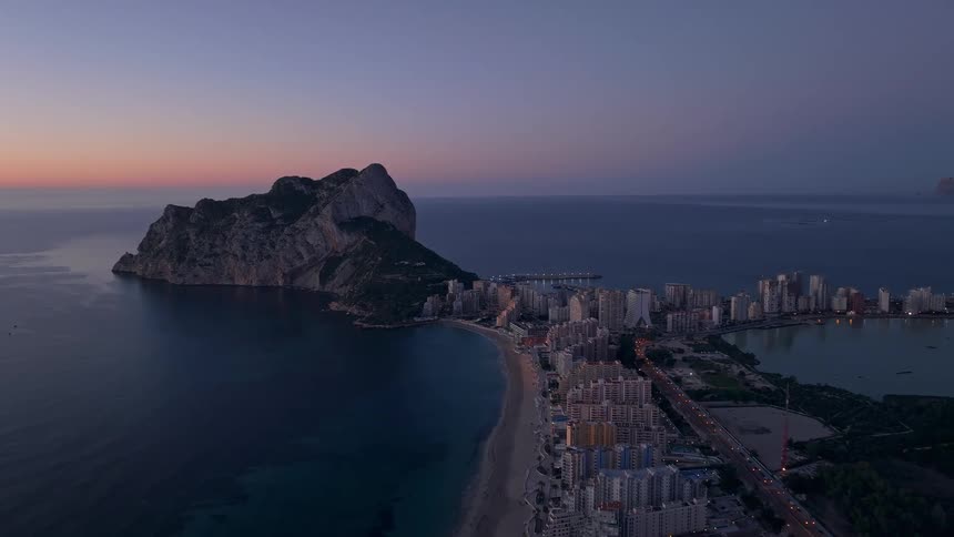 An aerial twilight view shows a massive rock formation looming over the dense skyline of a city as city lights begin to flicker along the curved sandy beach and the calm sea