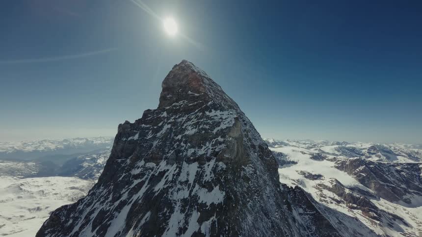 An aerial view shows a massive jagged mountain peak with steep rocky faces and patches of white snow under a bright sun and clear blue sky with a wide range of snowy mountains in the distance
