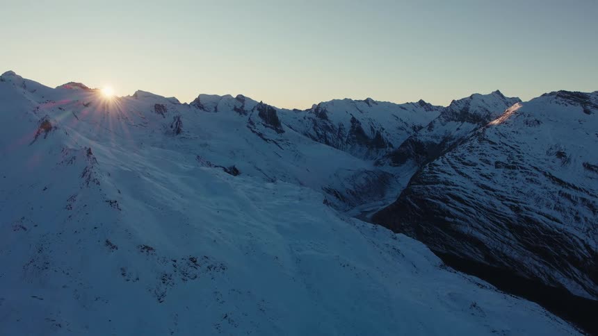 A wide aerial view shows jagged mountain peaks covered in thick white snow during a golden sunset as the sun peeks over a cold ridge and casts long shadows across the rugged alpine terrain under a clear sky