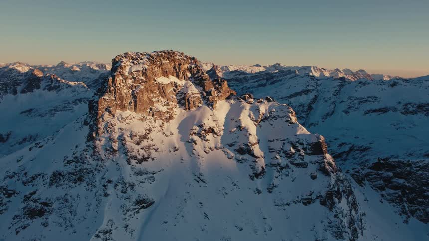 A wide aerial view shows a jagged rocky peak in the alps covered in white snow as the low sun of a winter morning casts a warm orange glow and long shadows across the rugged mountain range
