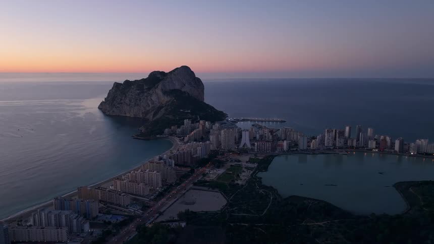 A wide aerial view shows a coastal city with high rise buildings and a large saltwater lake located next to a massive rock formation at the edge of the sea during a purple and orange sunrise