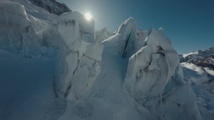 A drone flies over a massive blue and white glacier with deep cracks and jagged ice formations under a bright sun in a clear sky over a high mountain range