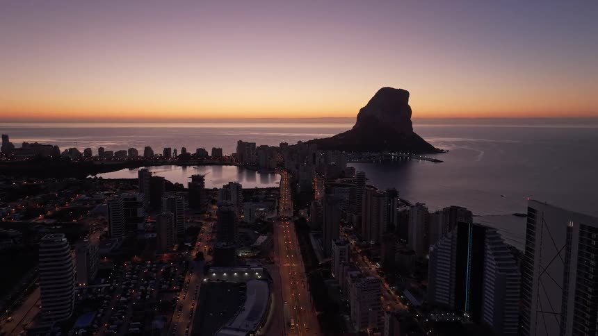 A coastal city during twilight where a massive limestone rock rises from the sea as the sun glows orange on the horizon and street lights illuminate the long boulevard below high rise buildings and a quiet lagoon