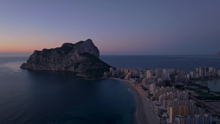 Coastal city with buildings along the beach and a large rock formation by the sea at dusk