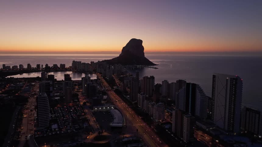 A wide aerial view shows the city of calpe at dawn with the peon de ifach landmark silhouetted against an orange horizon while streetlights and building windows glow across the urban landscape
