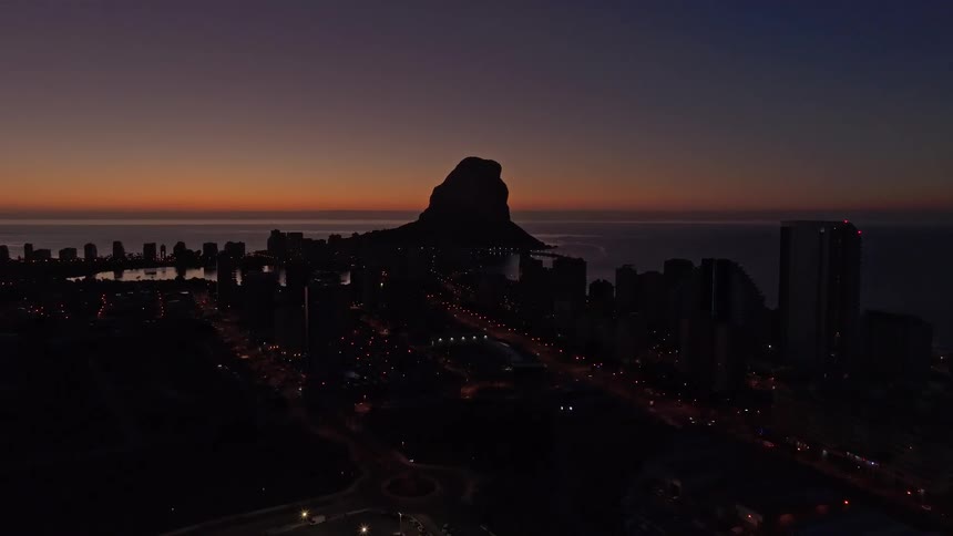 An aerial view of a coastal city skyline at dusk with a mountain silhouette on the horizon