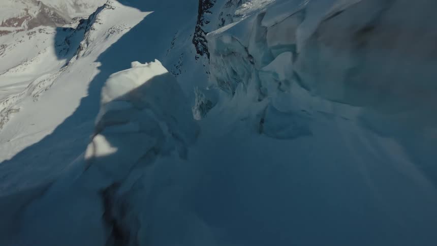 A fast aerial view dives over jagged blue ice formations and deep crevasses on a snow covered mountain glacier before banking sharply past a steep rocky cliff