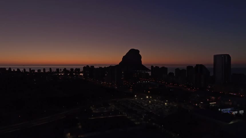 Silhouette of a coastal city at sunset with a mountain in the background and city lights on
