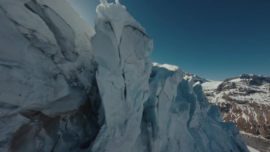 The camera glides closely alongside a massive jagged ice wall of a glacier eventually cresting over a snowy ridge to reveal the suns bright glare over a vast frozen mountain landscape