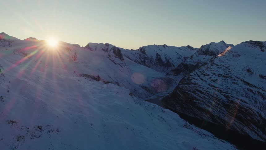 An aerial drone camera glides over a vast range of jagged mountain peaks covered in thick white snow while the morning sun peeks over a distant ridge creating a bright lens flare against a clear blue sky during the cold winter season