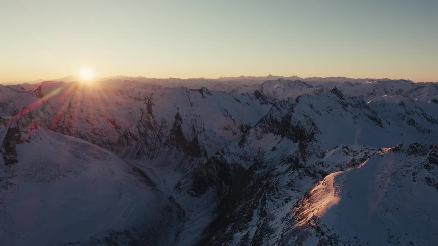 Snow covered rough rocky mountain terrain across the landscape as the sun shines brightly on the horizon