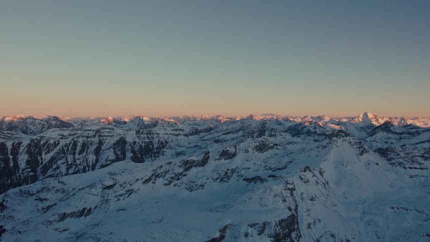 A wide aerial view shows the snowy peaks of the alps under a clear sky during a cold winter evening as the setting sun casts a soft orange light over the mountain range