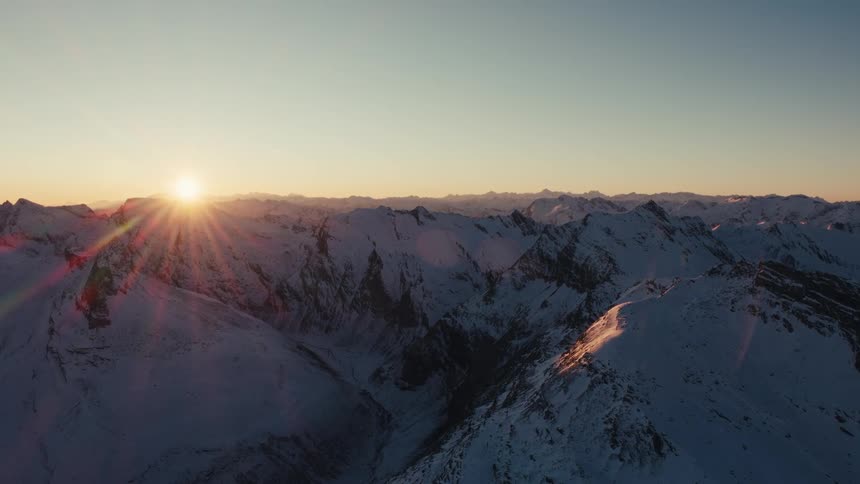 The sun sets over a vast range of jagged snowy mountains during golden hour as the orange light catches the peaks and casts long blue shadows across the frozen alpine valleys under a clear sky