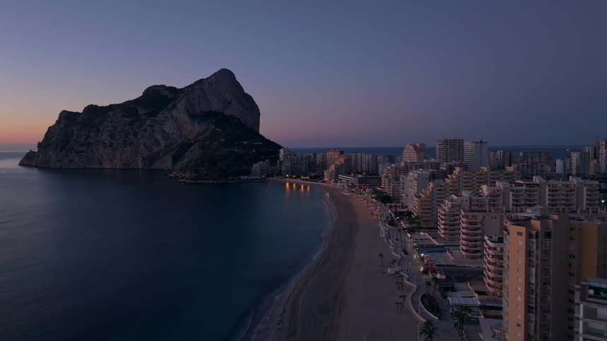 An aerial view shows the peon de ifach limestone rock towering over the city of calpe and its sandy beach during a purple sunrise while city lights reflect on the calm sea water