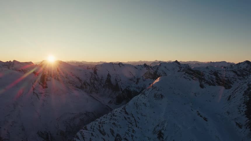 Snow covered rough rocky mountain terrain across the landscape as the sun shines brightly on the horizon