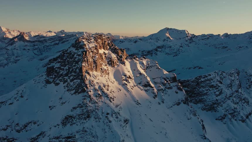 Sunlight shines on the rough terrain of a mountain peak with snow covered mountains in the distance