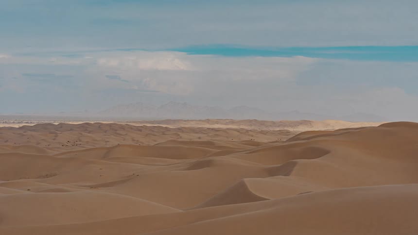 A wide view of sand dunes shows rolling tan sand dunes stretching toward a horizon with distant mountains under a blue sky with white clouds