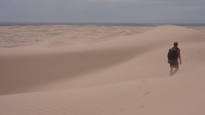 Man with a backpack walks across sand dunes in a desert landscape