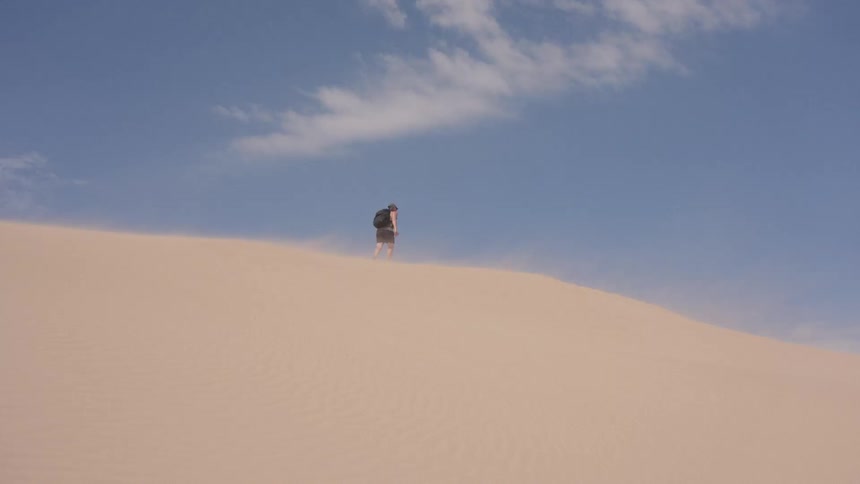 A person wearing a backpack walks along the ridge of a large tan sand dune while wind blows sand across the surface under a blue sky with light clouds