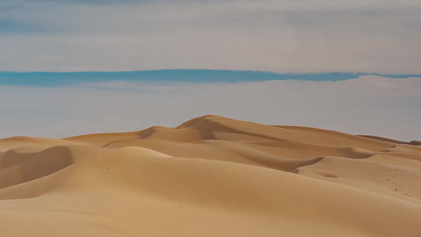 A wide view shows rolling tan sand dunes under a blue sky with soft white clouds in a vast desert landscape