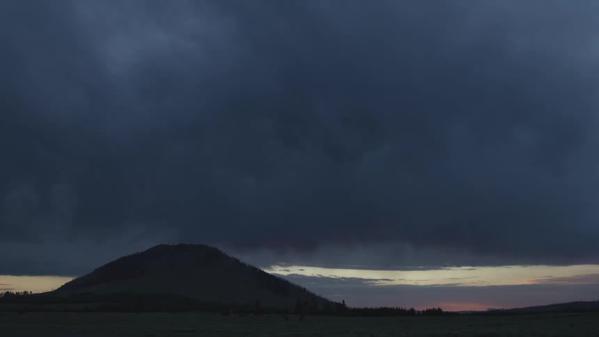 A wide view shows a dark mountain peak silhouetted against a deep blue dusk sky as thick heavy storm clouds move across the horizon where a faint orange light remains