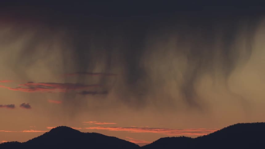 Rain curtains descend from dark clouds over mountain silhouettes at sunset