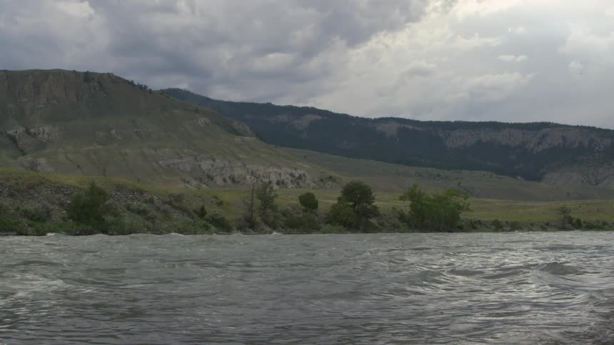 A wide river flows through a rugged valley with grassy hills and scattered trees beneath a cloudy sky
