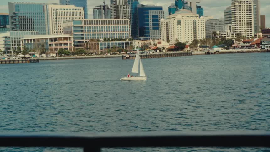A wide shot shows a small white sailboat moving across a body of water with a city skyline and tall buildings in the background under a bright sky