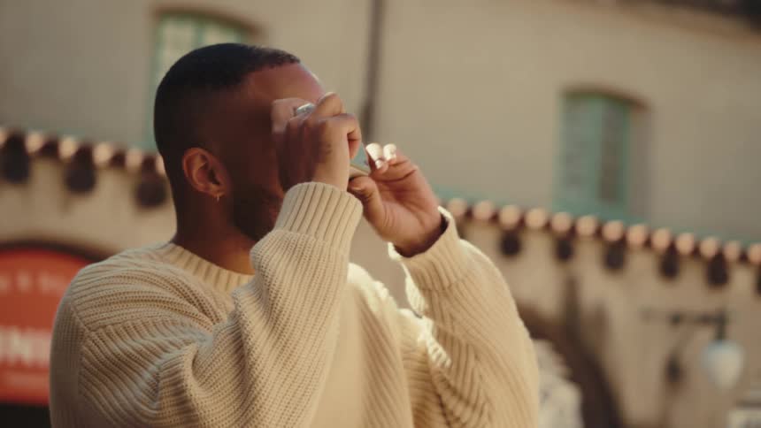 A medium shot shows a man wearing a cream sweater as he stands outdoors and uses a camera to take a picture