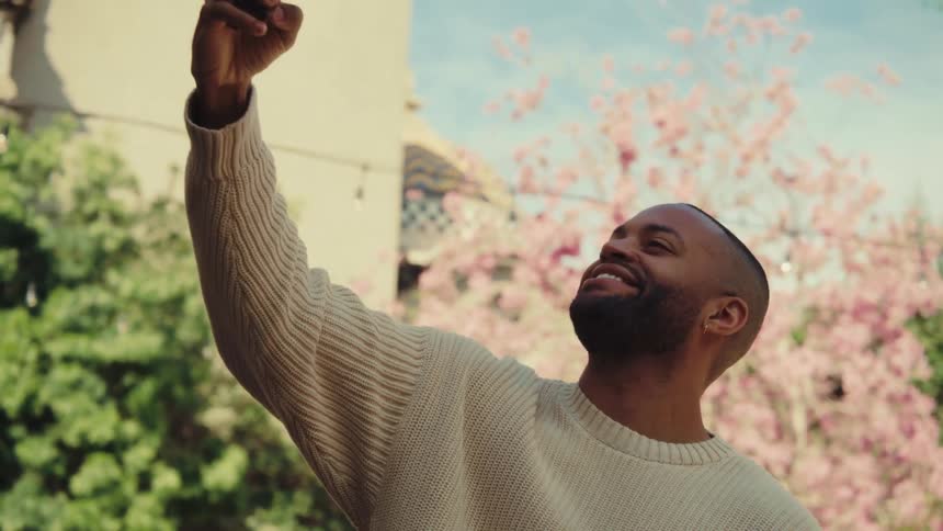A medium shot shows a man standing outdoors in a cream sweater as he looks at his phone before raising it to take a selfie with a smile against a backdrop of green trees and pink blossoms