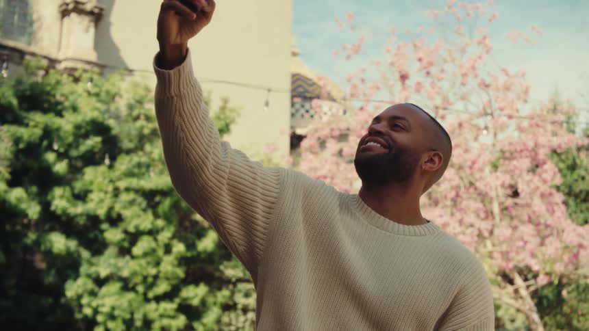 A medium shot shows a man standing outdoors in a cream sweater as he looks at his phone before raising it to take a selfie with a smile against a backdrop of green trees and pink blossoms
