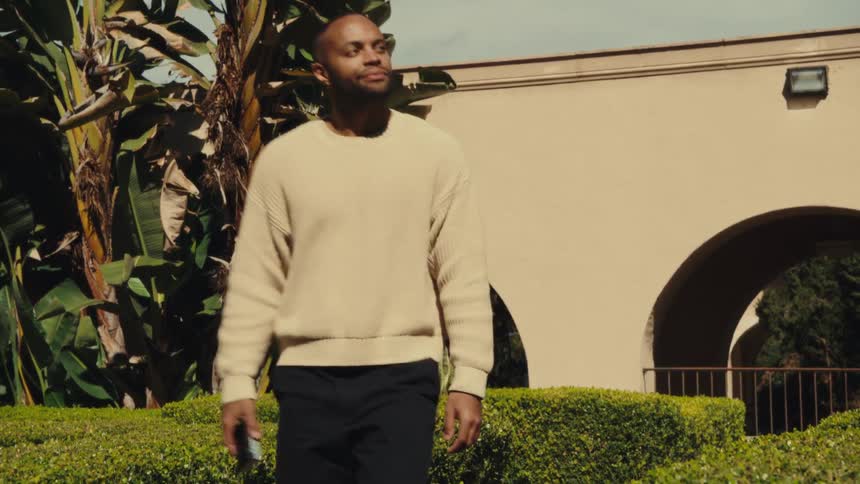 A man in a cream sweater walks holding a smart phone confidently through a sunny courtyard lined with greenery and arched architecture
