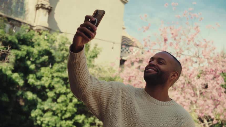 A medium shot shows a man standing outdoors in a cream sweater as he looks at his phone before raising it to take a selfie with a smile against a backdrop of green trees and pink blossoms