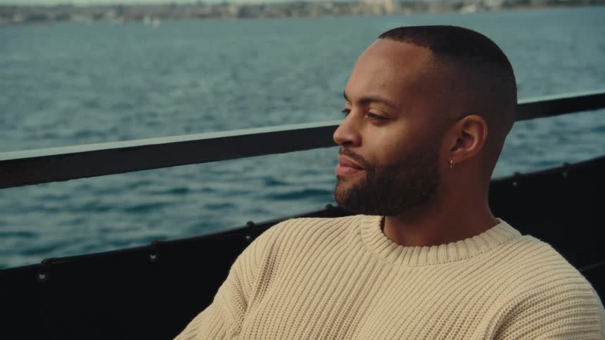 A man in a beige sweater sits by the water looking thoughtfully toward the distance