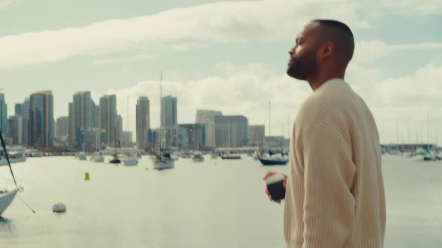 A black man in a cream sweater stands by a harbor holding a coffee cup while looking at a city skyline and several sailboats anchored in the water under a cloudy sky