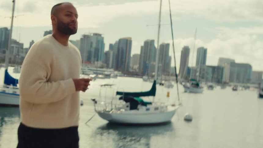A man in a beige sweater holds a drink while standing by a harbor with sailboats and a city skyline in the background