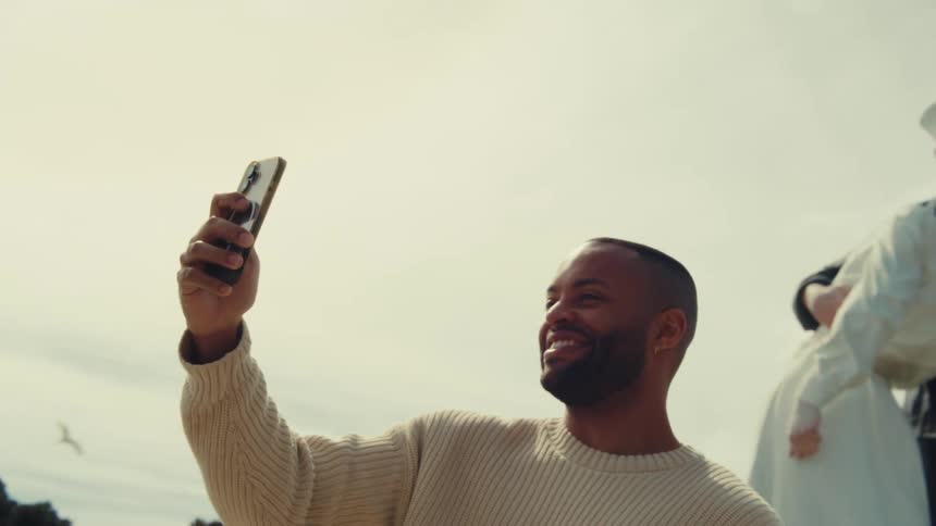 A man wearing a cream sweater uses his phone to take a selfie in front of a large statue depicting a sailor kissing a woman in a white dress under a bright sky
